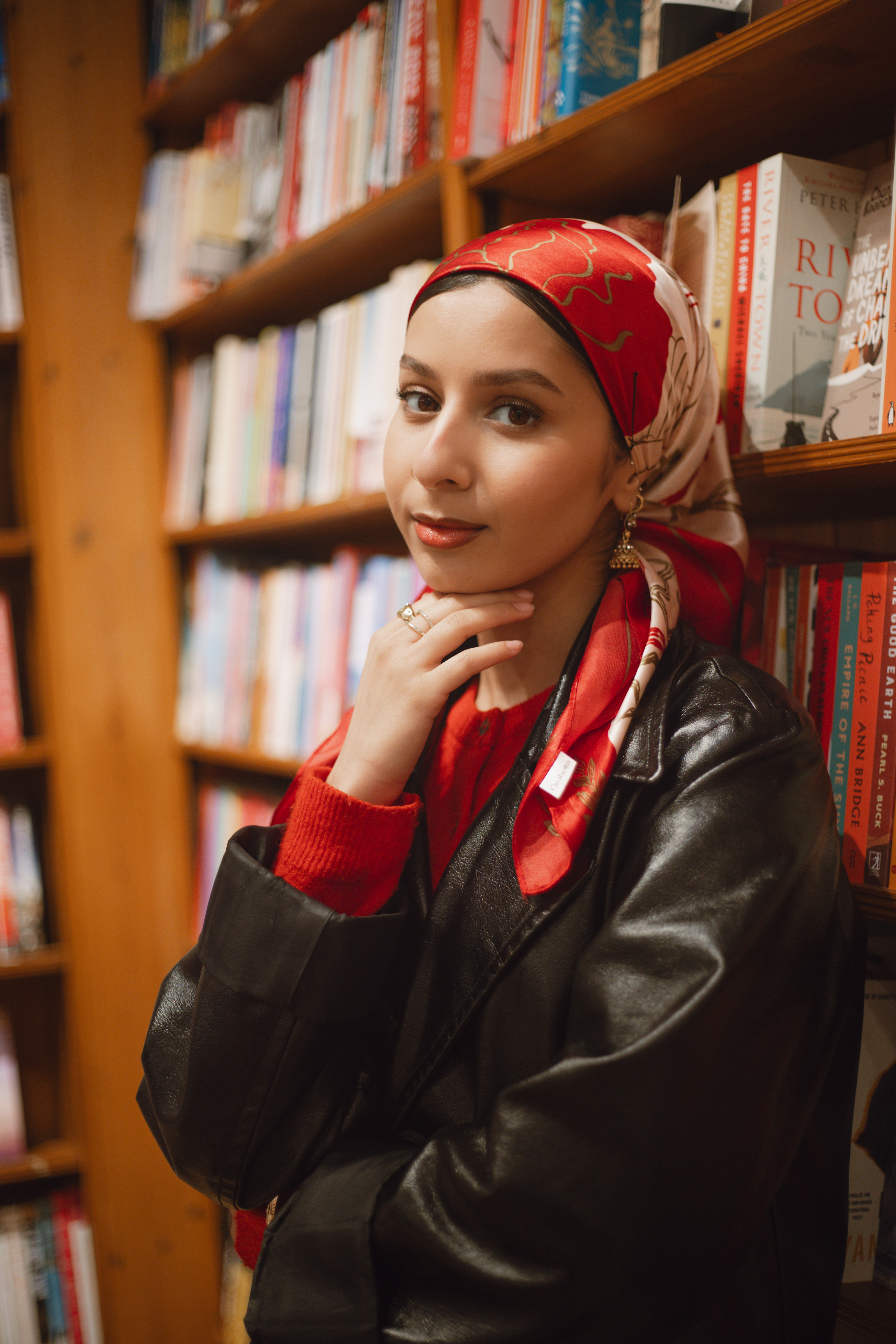 Furvah Shah, photographed in a London bookstore, wearing a red headscarf and cardigan against a wall of books.